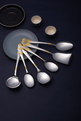 Seven-piece serving spoon set with gold-embellished handles fanned over blue-black table setting with plates.