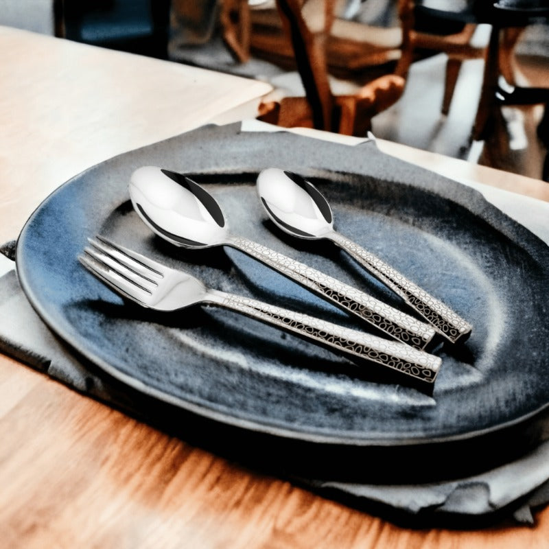 Three-piece patterned cutlery set placed on a dark ceramic plate with soft napkin and wooden table.