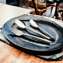 Three-piece patterned cutlery set placed on a dark ceramic plate with soft napkin and wooden table.