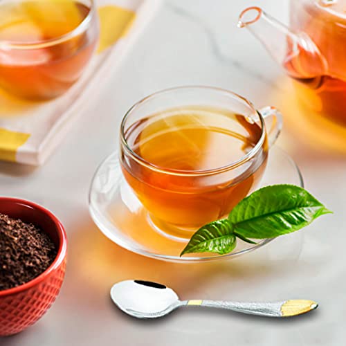 Glass teacup with fresh tea leaves, red bowl of loose tea, and silver-gold teaspoon placed nearby.