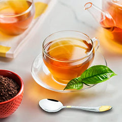 Glass teacup with fresh tea leaves, red bowl of loose tea, and silver-gold teaspoon placed nearby.