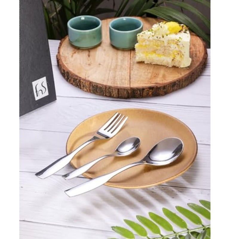 Modern stainless steel cutlery set on ceramic plate, styled with dessert, cups, and wooden serving board.