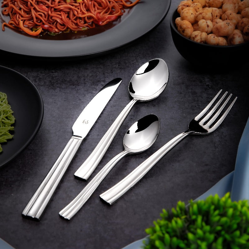 Glossy stainless steel cutlery set placed beside plates of noodles, chickpeas, and herbs on black tabletop.