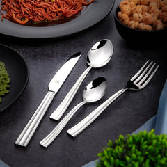 Glossy stainless steel cutlery set placed beside plates of noodles, chickpeas, and herbs on black tabletop.