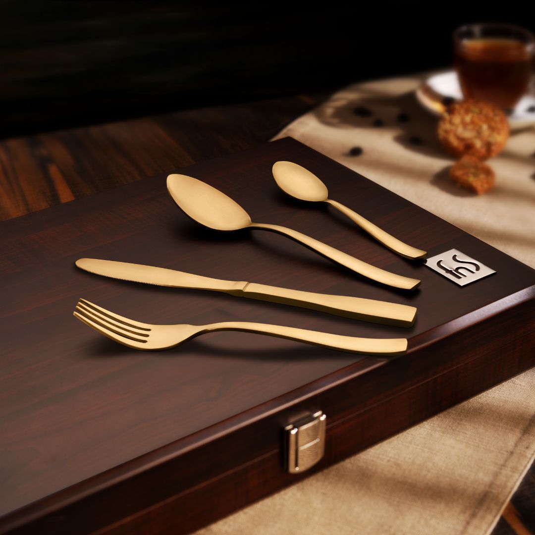 Refined gold flatware arranged on a wooden box lid, styled with coffee and biscuits in background.