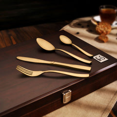 Refined gold flatware arranged on a wooden box lid, styled with coffee and biscuits in background.