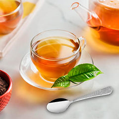 Small spoon with textured handle beside glass teacup, teapot, and tea leaves on marble surface.