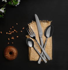 Elegant cutlery set placed on burlap fabric beside chocolate donut and scattered chips on black surface.