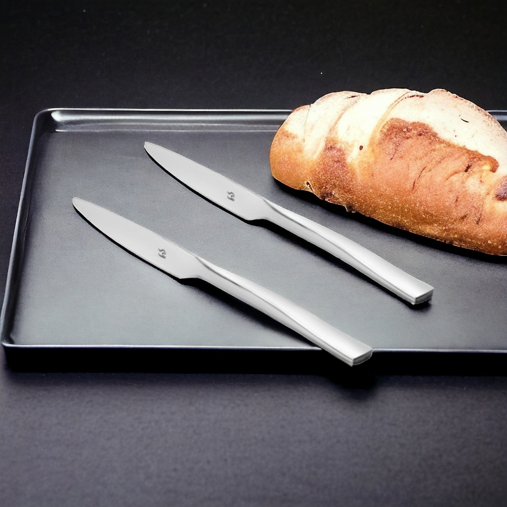Two stainless steel dinner knives placed on dark baking tray beside freshly baked artisan bread loaf against dramatic black background.