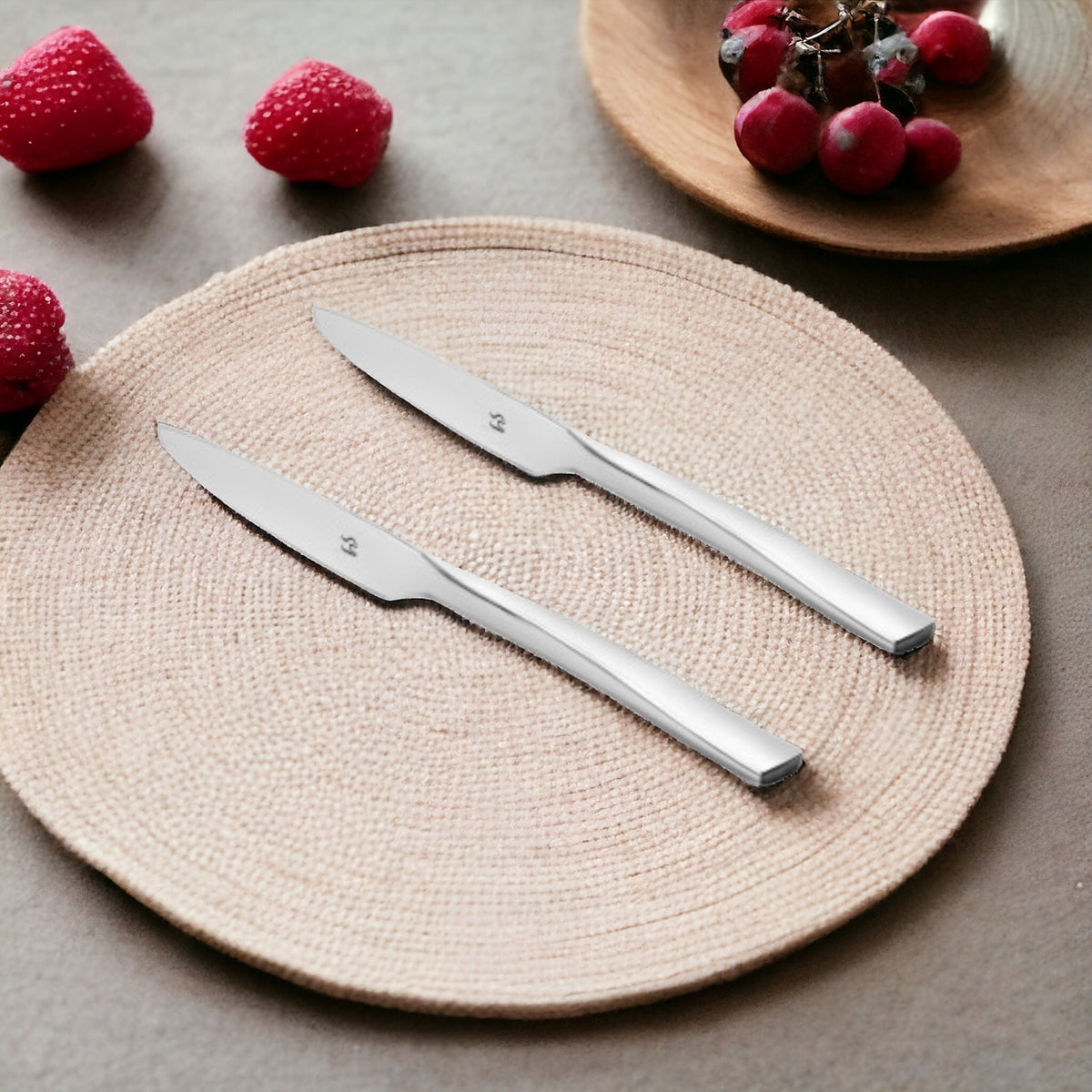 Two sleek stainless steel dinner knives on beige woven placemat with fresh strawberries and red grapes in background setting.