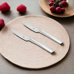Two sleek stainless steel dinner knives on beige woven placemat with fresh strawberries and red grapes in background setting.