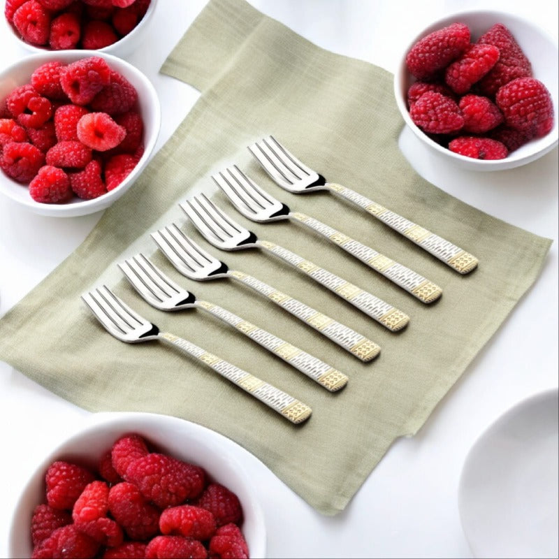 A set of six decorative stainless steel dinner forks with gold accents placed neatly on a light green napkin, surrounded by bowls of fresh raspberries.