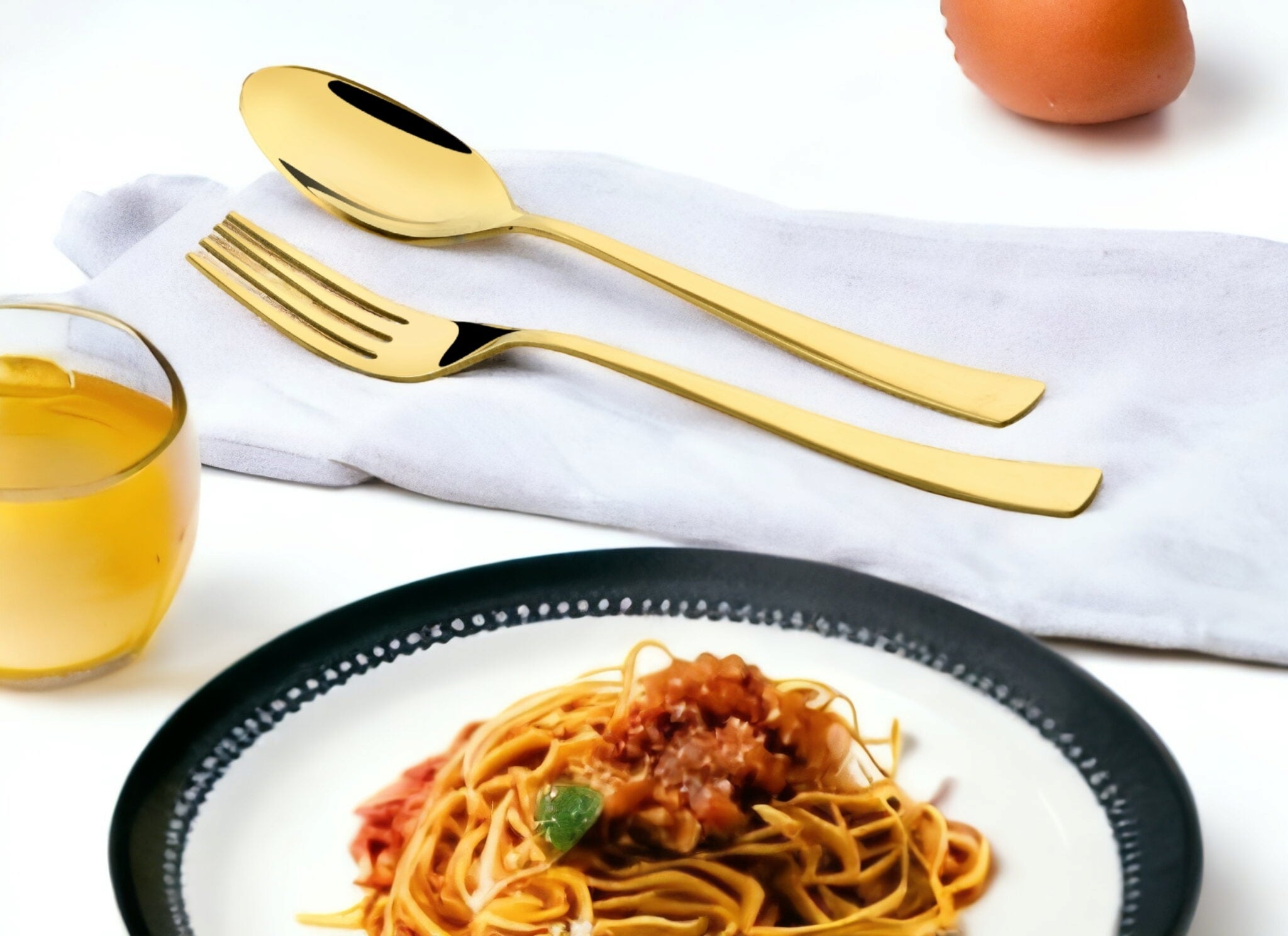 Elegant gold cutlery placed on napkin next to plate of spaghetti and glass of juice.
