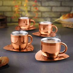 Set of 4 fns gold finish stainless steel tea cups with matching square saucers filled with steaming hot tea, styled on a grey surface with cookies and autumn foliage in the background