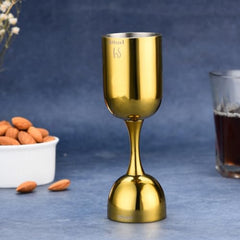 FNS gold plated double jigger with 60ml and 30ml measurements on blue surface with white bowl of almonds and dark beverage glass