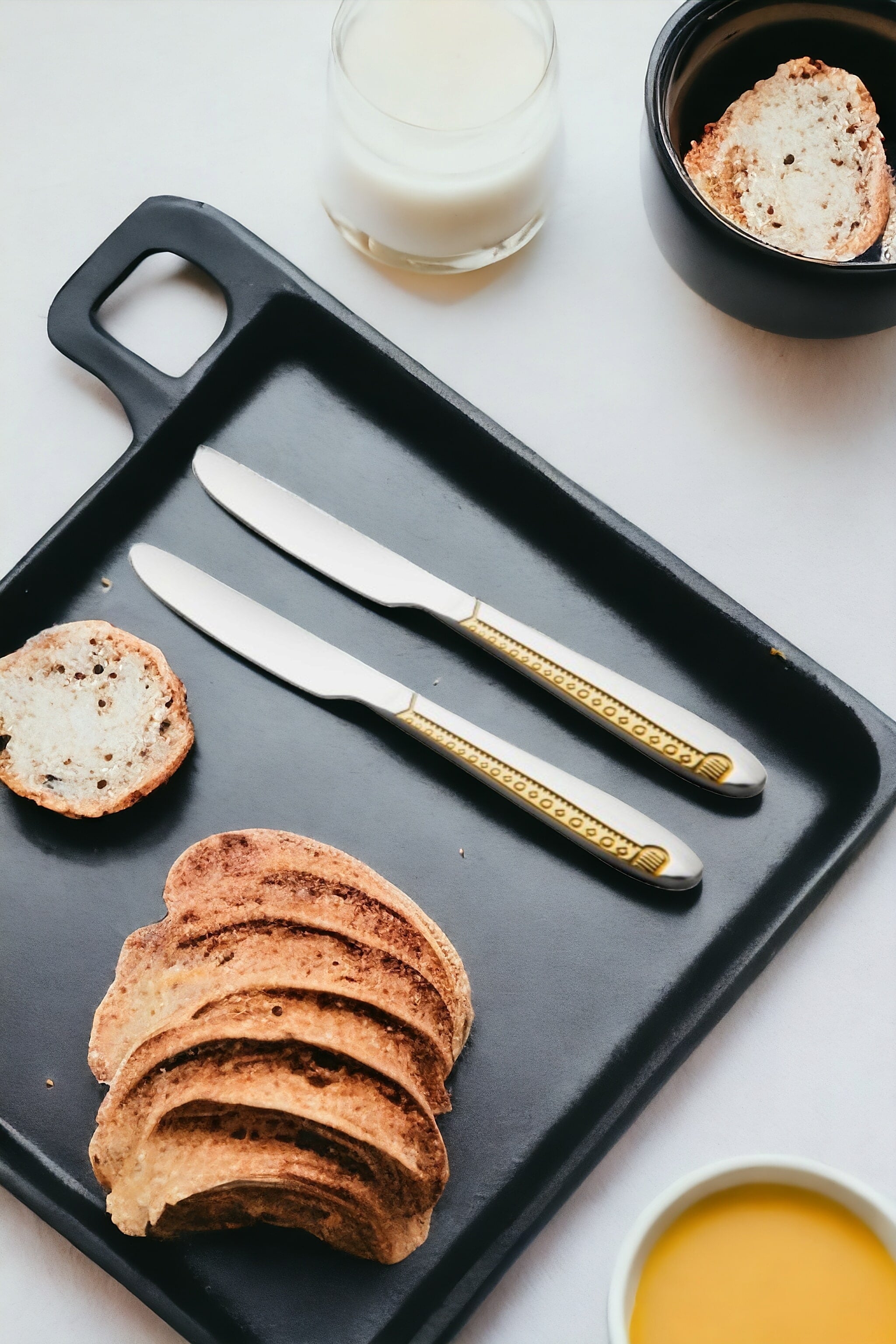 Two luxury dinner knives with gold-plated handles on black serving board with sliced artisan bread, orange juice, and milk for breakfast.