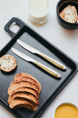 Two luxury dinner knives with gold-plated handles on black serving board with sliced artisan bread, orange juice, and milk for breakfast.