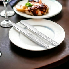 Two stainless steel Helios dinner knives with textured handles placed on white plate beside wine glass and gourmet meal setting.
