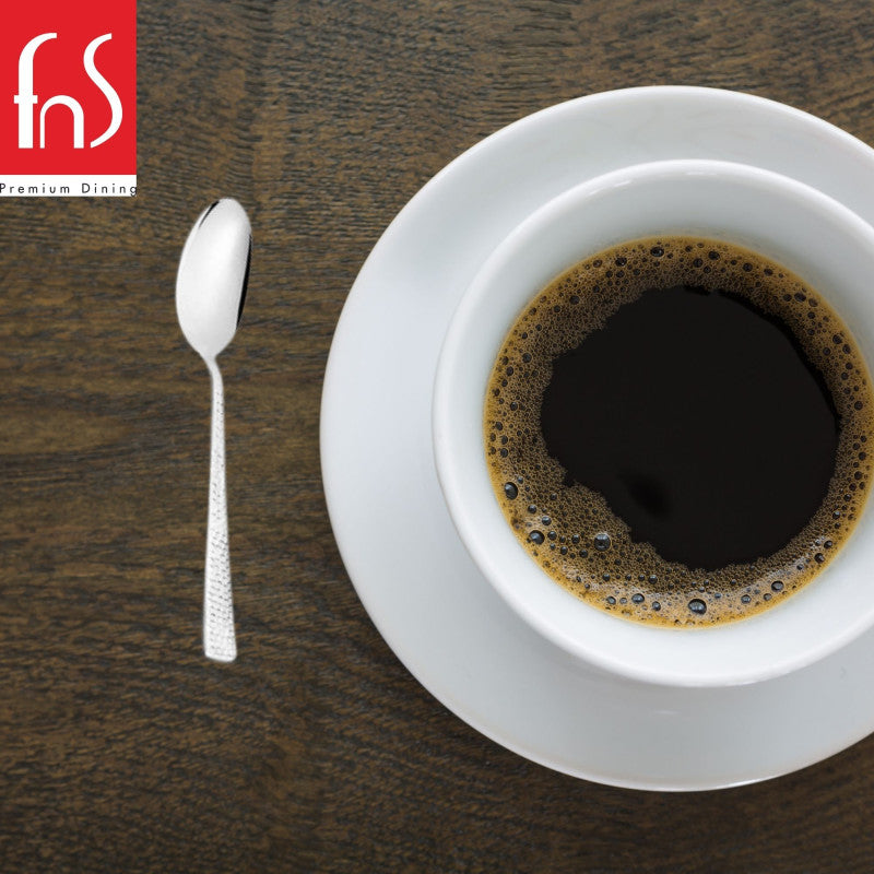 Helios teaspoon placed beside white coffee cup and saucer on wooden table, showcasing elegant mirror finish and textured handle in lifestyle setting.