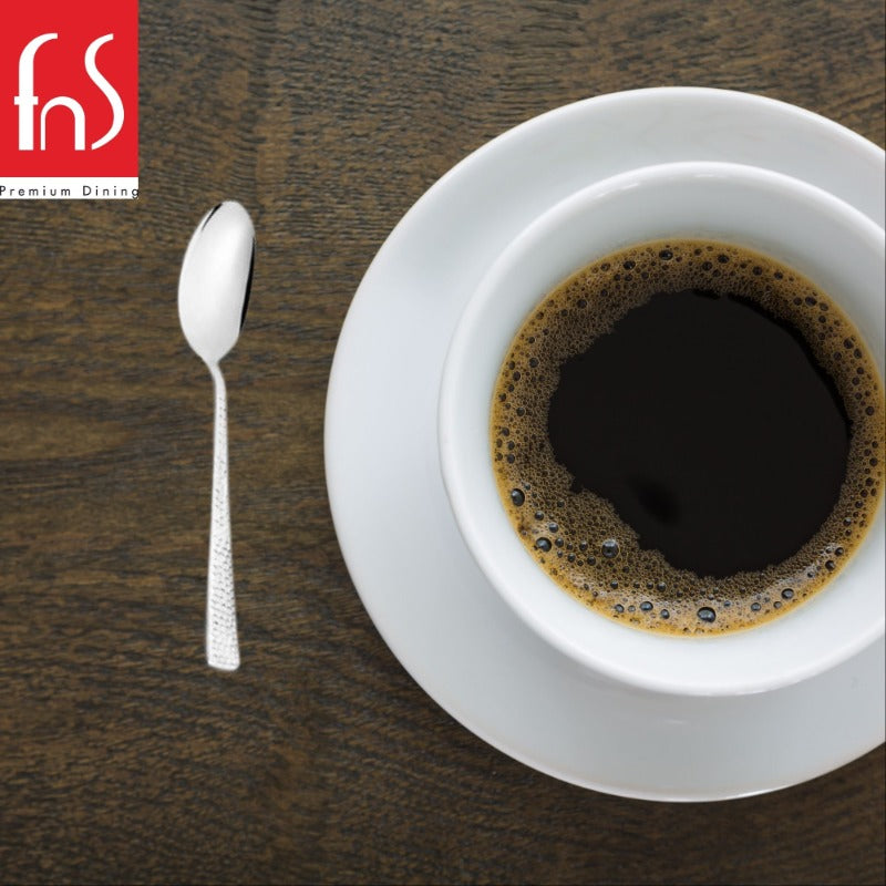 Stainless steel teaspoon with textured handle placed beside white coffee cup and saucer on dark wooden table in minimalist composition.