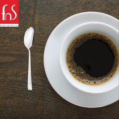 Stainless steel teaspoon with textured handle placed beside white coffee cup and saucer on dark wooden table in minimalist composition.