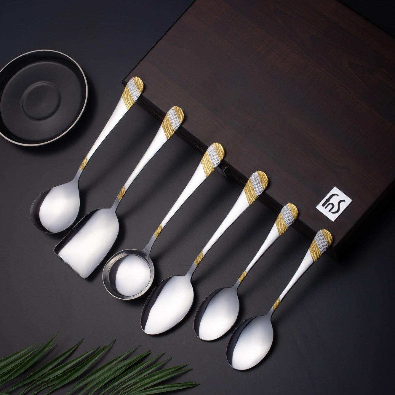 FNS Imperio 24k gold plated stainless steel 6-piece serving set arranged on brown leatherette box with dark background and palm leaf accent