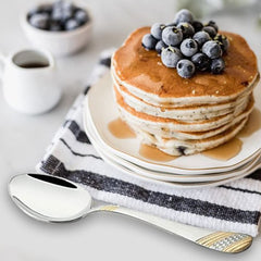 FNS Imperio 24-karat gold-plated large serving spoon displayed with blueberry pancake stack, coffee cup, and striped napkin creating elegant breakfast table lifestyle setting.