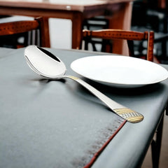FNS Imperio large serving spoon with gold-plated textured handle positioned on dining table with white plate and restaurant chairs in blurred background setting.
