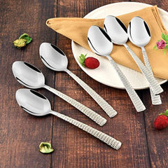 Six stainless steel spoons with textured handles arranged on a wooden table with yellow napkins, white plates, and fresh strawberries as decorative elements