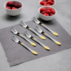 Six stainless steel dinner forks with gold-colored tips laid out on a gray cloth napkin. Two white bowls filled with red and dark berries are in the background.