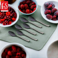 Six black teaspoons with decorative silver geometric handles arranged on green mat, surrounded by white bowls filled with mixed berries and raspberries.