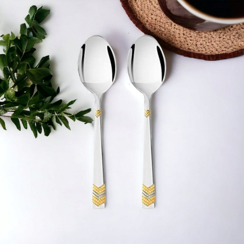Six stainless steel teaspoons with gold chevron pattern details on white rectangular plate, surrounded by coffee cups and chocolate cake on gray surface