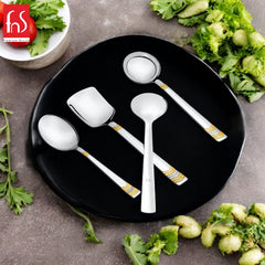 FNS Raga 4-piece gold accent serving utensil set arranged on black plate with fresh greens and vegetables overhead table setting view