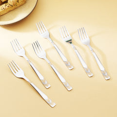 Six stainless steel fruit forks scattered on beige surface near pastries, displaying chevron gold pattern design details