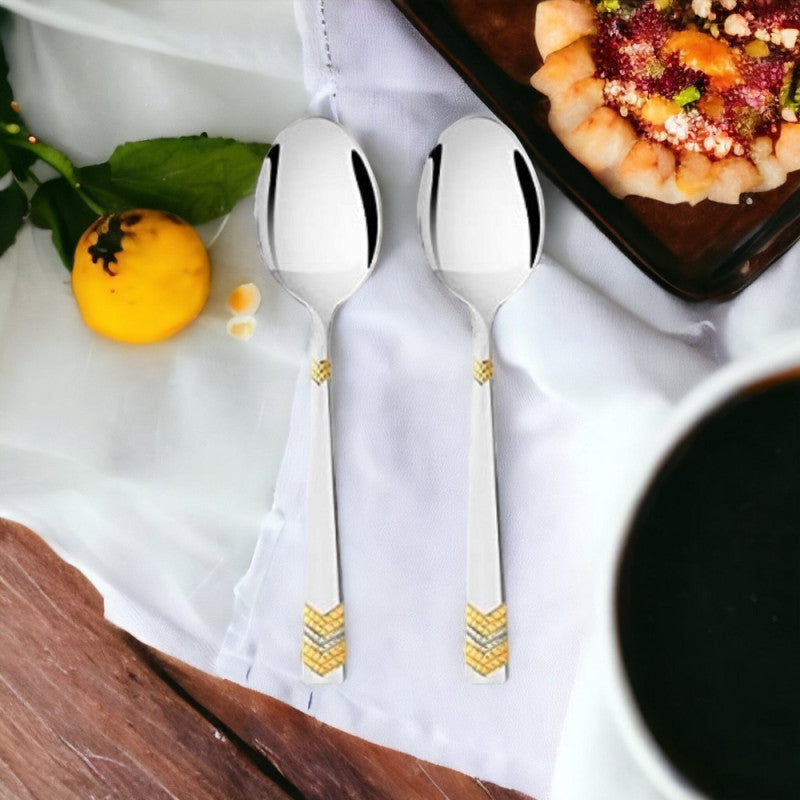 Two FNS Raga gold plated dinner spoons with chevron pattern handles displayed on white napkin beside dessert bowl and coffee on wooden table
