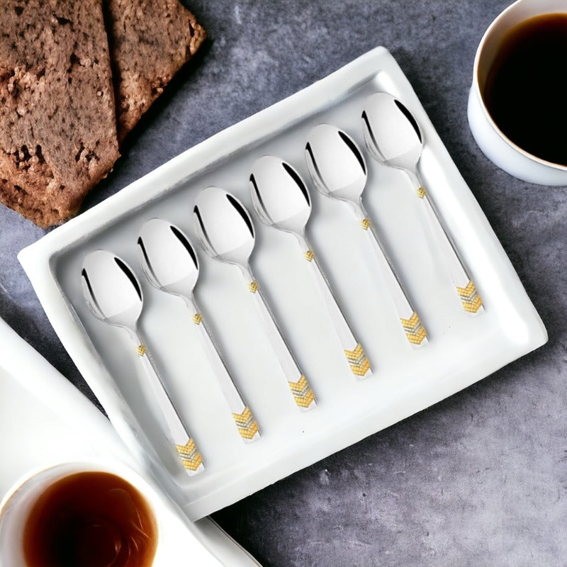 Six stainless steel teaspoons with gold chevron pattern details on white rectangular plate, surrounded by coffee cups and chocolate cake on gray surface