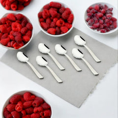 Six stainless steel teaspoons with textured handles arranged on a gray surface, surrounded by white bowls filled with fresh red and purple raspberries.