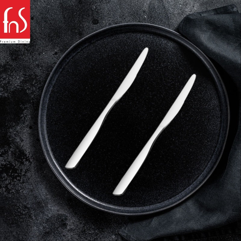Two FnS stainless steel Rio dinner knives arranged parallel on round black plate with dark textured background and fabric napkin.