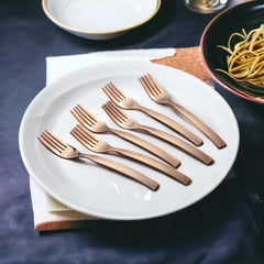 Six copper-colored forks arranged on white plate with pasta bowl nearby