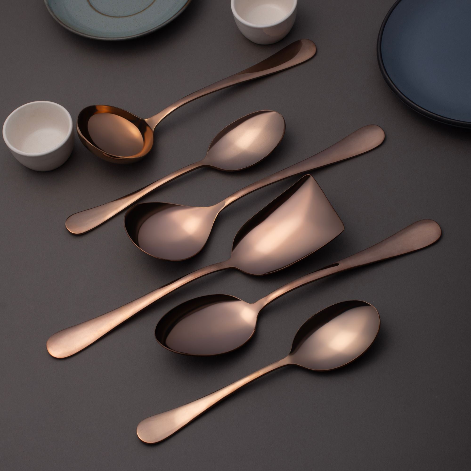 FNS Rosella rose gold 6-piece serving utensil set lifestyle display with ceramic plates, bowls, and modern table setting on dark gray surface