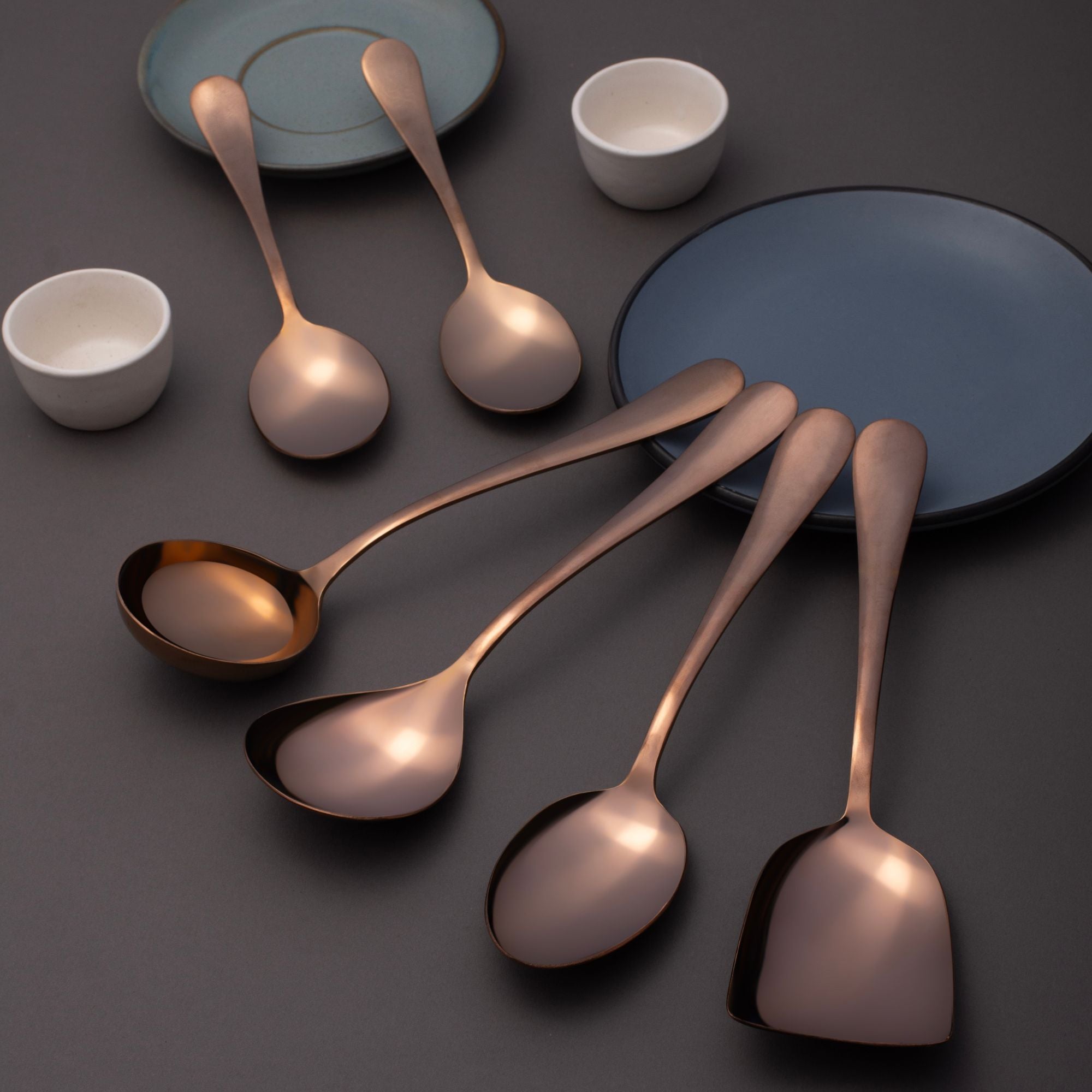 FNS Rosella rose gold 6-piece serving spoon set with mirror finish displayed on dark gray surface with blue ceramic plates and bowls