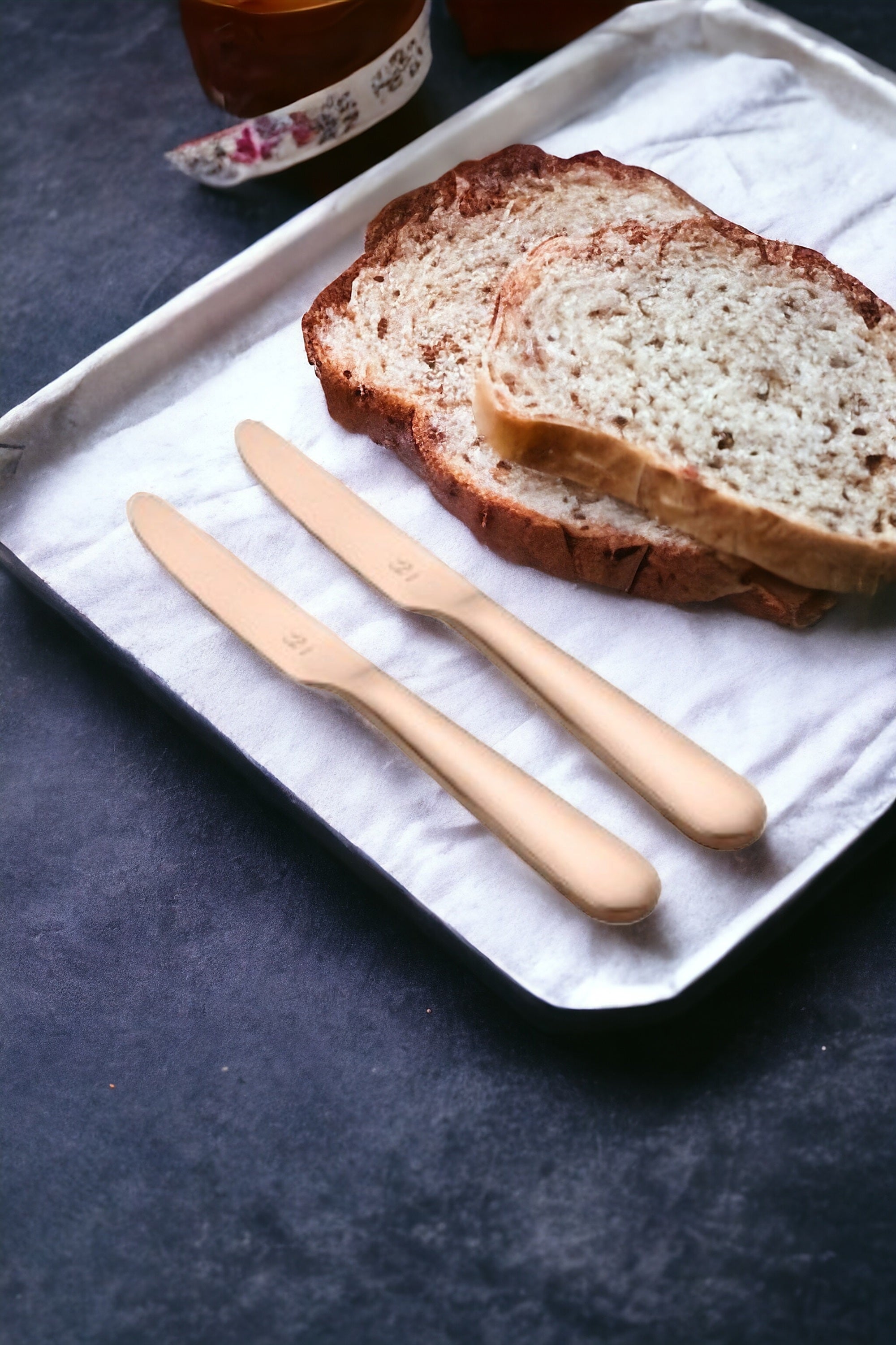Two rose gold dinner knives displayed on white napkin with sliced artisan bread and breakfast beverages in elegant morning table setting.