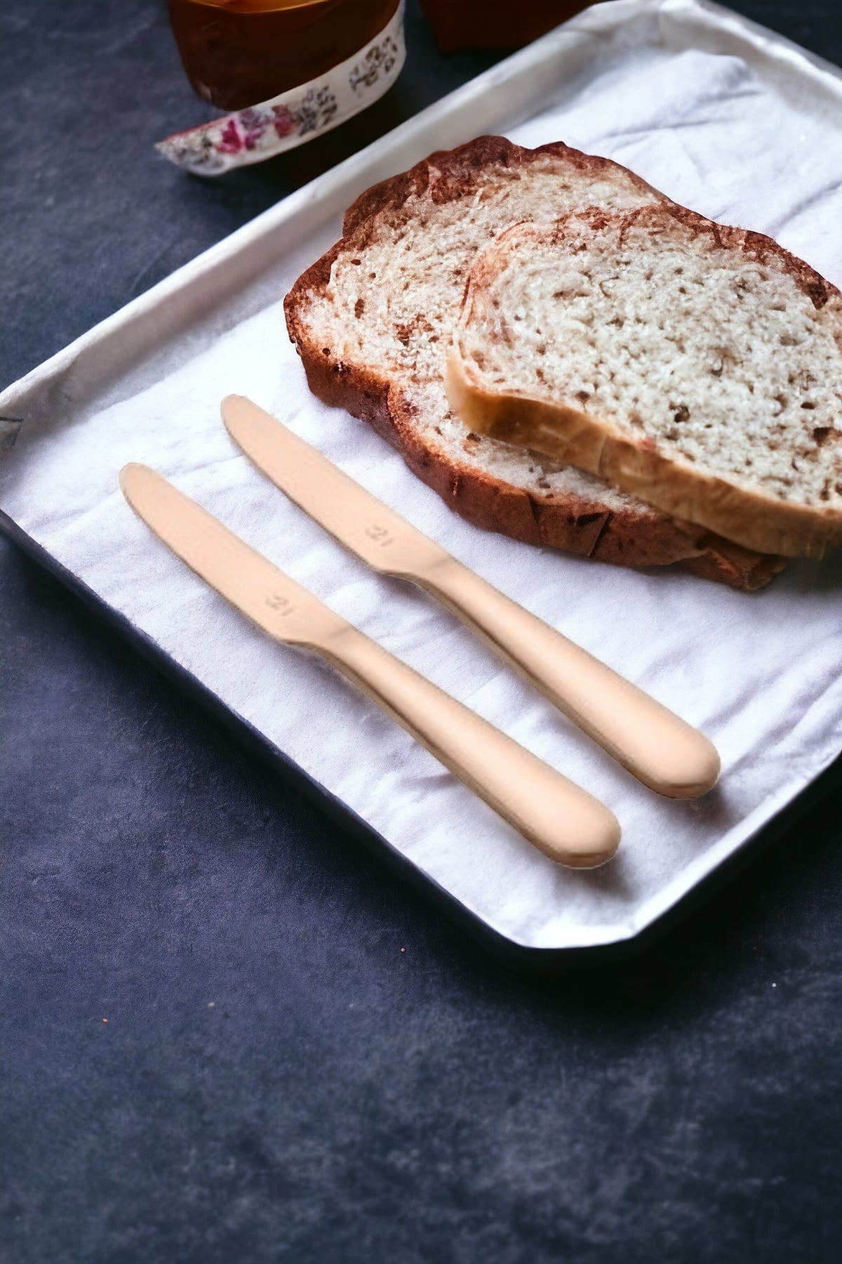 Two rose gold dinner knives displayed on white napkin with sliced artisan bread and breakfast beverages in elegant morning table setting.