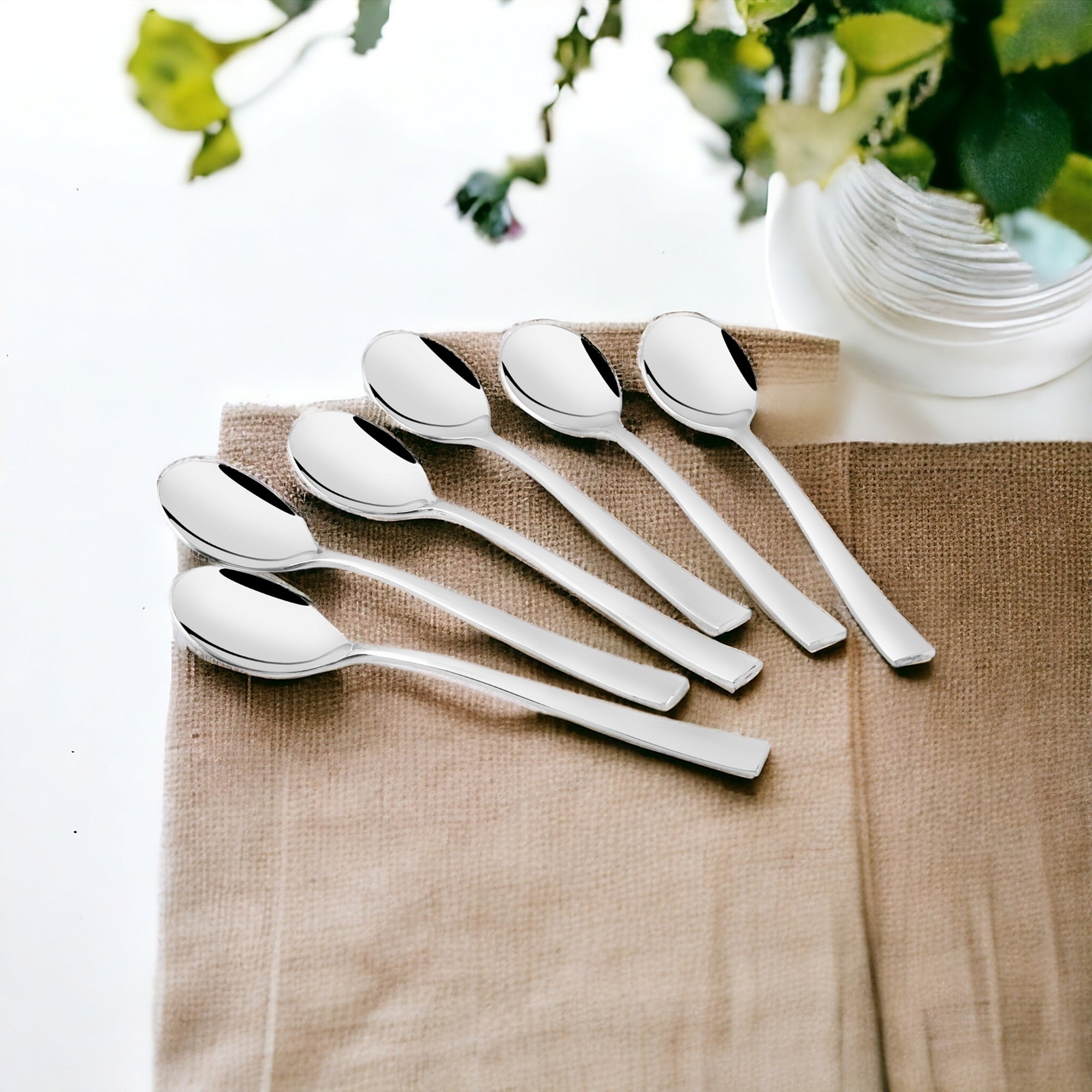Six stainless steel spoons with flat modern handles arranged on beige burlap placemat with white ribbed bowl and greenery in background for elegant table setting