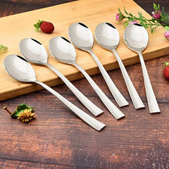 Six mirror-polished stainless steel teaspoons with flat tapered handles arranged on wooden cutting board with strawberries and pink flowers on rustic surface