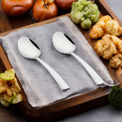 FNS Solo 2-piece serving spoon set displayed on wooden tray with gray napkin, fresh apples, cauliflower, and fried snacks creating rustic kitchen lifestyle scene.