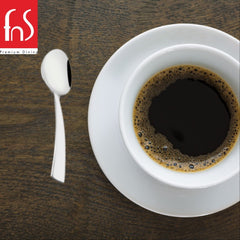 Single mirror-polished stainless steel teaspoon with angled flat handle beside white cup of black coffee on dark wooden surface, FnS Premium Dining logo