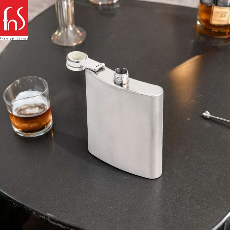 FNS stainless steel hip flask with integrated shot cup cap on dark bar counter with whiskey glass and bar tools