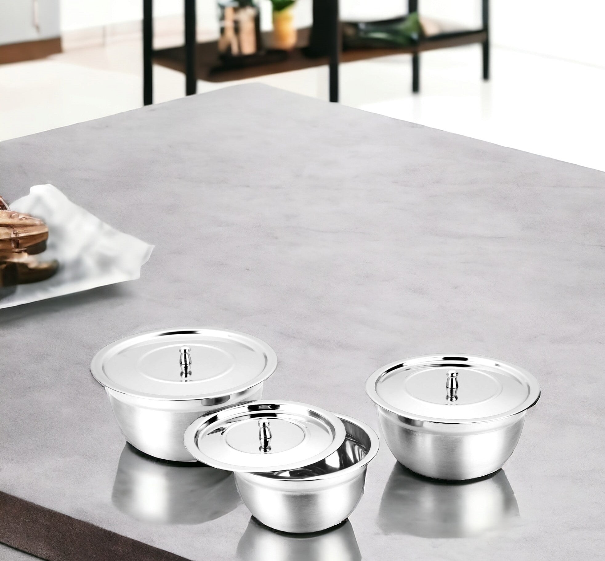 FNS stainless steel serving bowl set of 3 with steel lids arranged on a grey marble kitchen counter, with a modern open shelving unit and wooden cutting board in the background