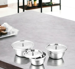 FNS stainless steel serving bowl set of 3 with steel lids arranged on a grey marble kitchen counter, with a modern open shelving unit and wooden cutting board in the background